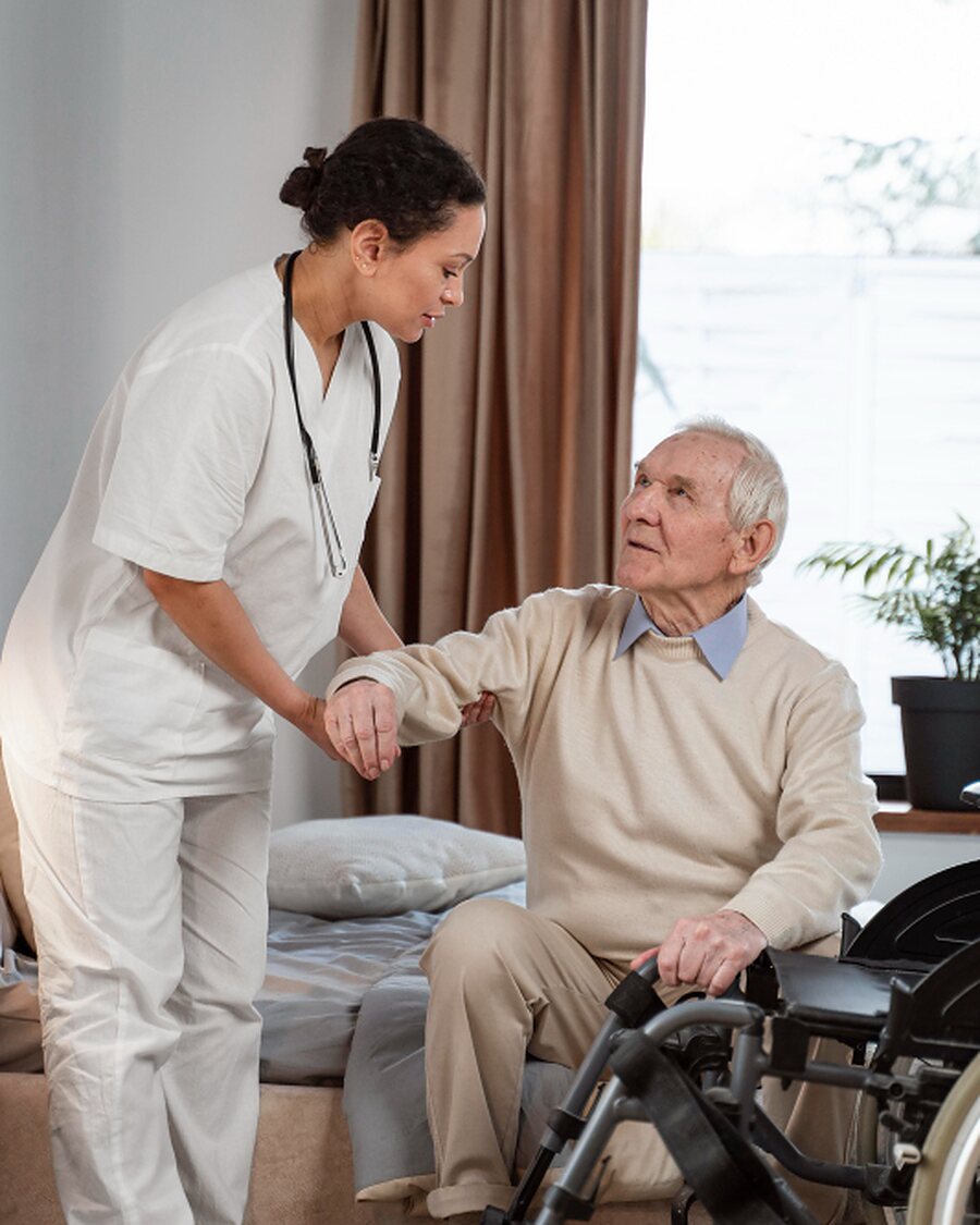 A caregiver supporting an older man as he sits at the edge of his bed
