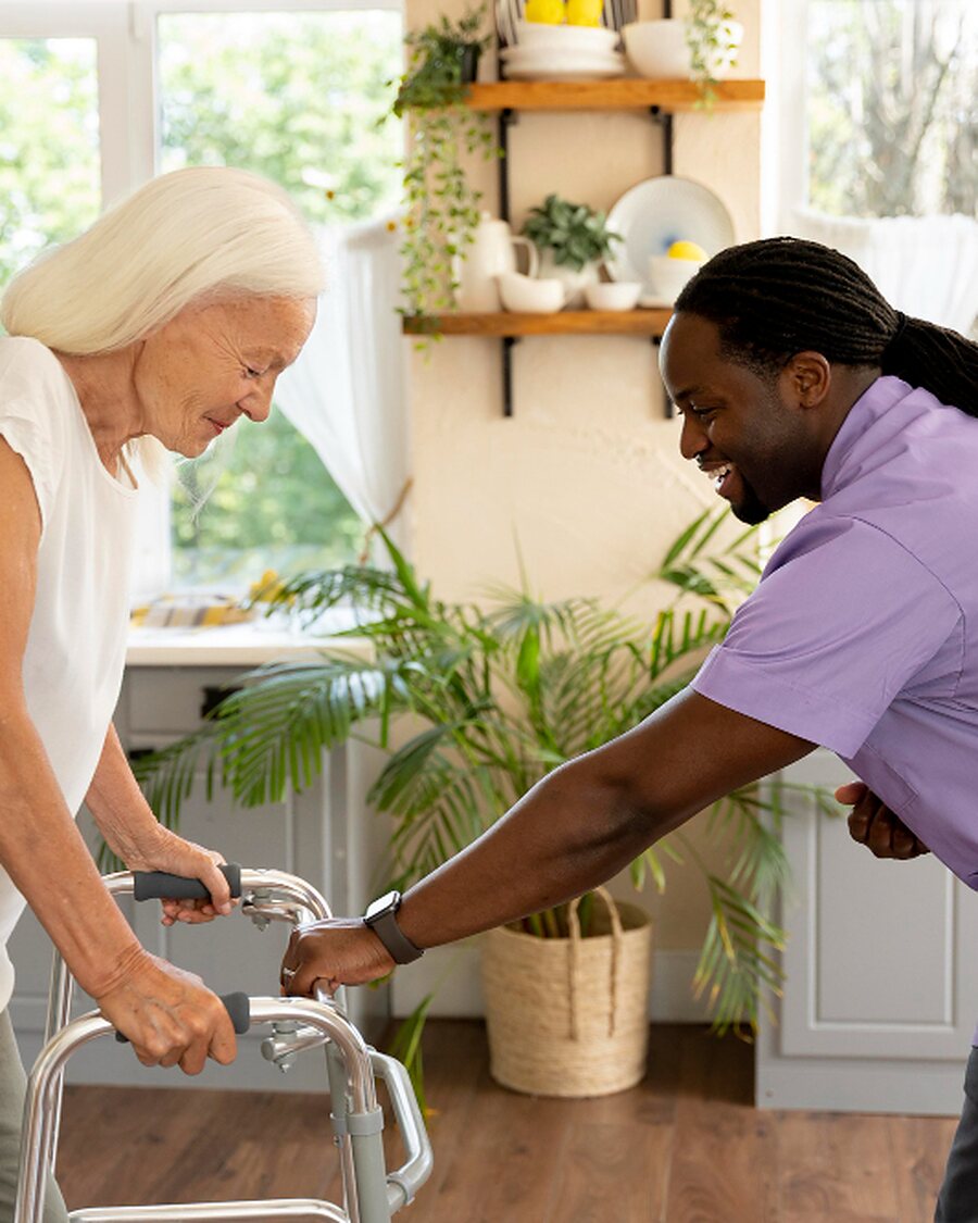 A caregiver steadying an older woman as she walks with her rollator in her kitchen